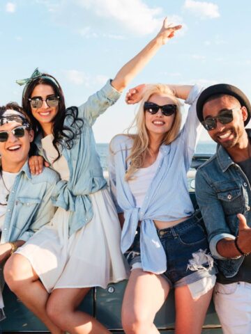 Four friends in casual summer clothes pose and smile by a blue car near the ocean on a sunny day.
