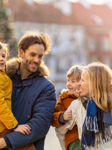 A family of four in winter coats smiles and walks together outdoors in a sunlit town square.