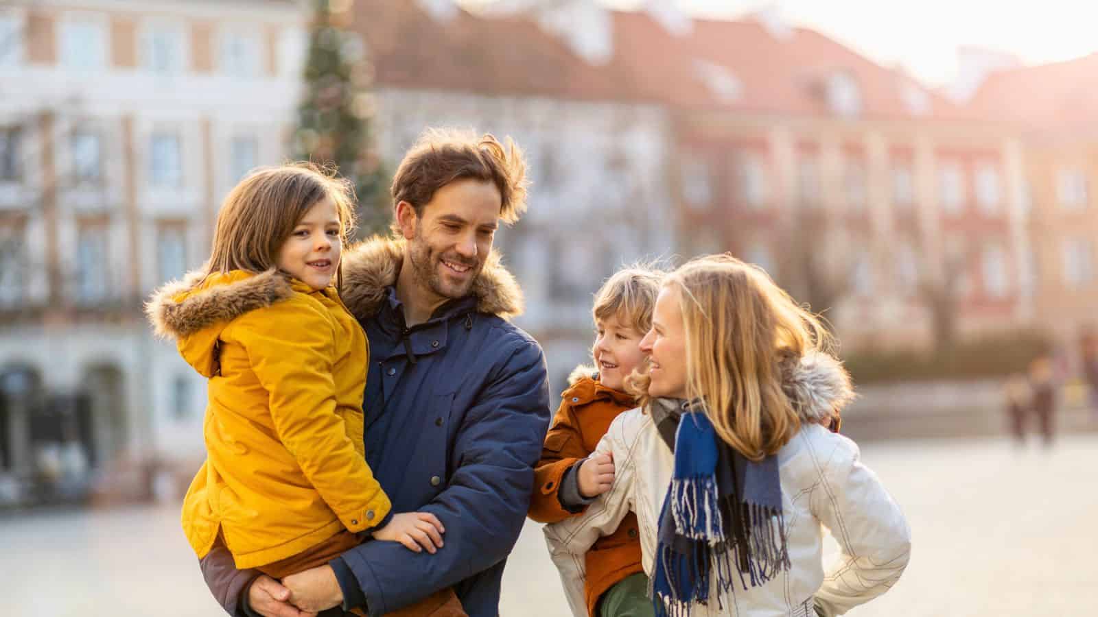 A family of four in winter coats smiles and walks together outdoors in a sunlit town square.