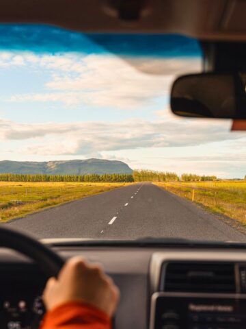 View from inside a car driving on an empty rural road toward distant green fields and mountains under a blue sky.
