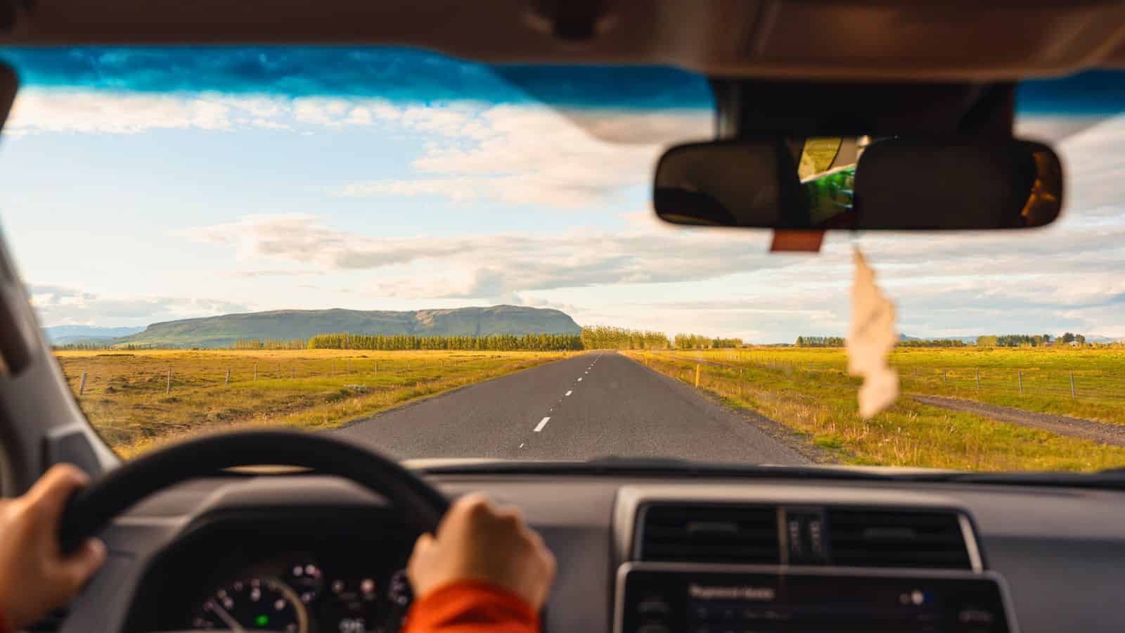 View from inside a car driving on an empty rural road toward distant green fields and mountains under a blue sky.