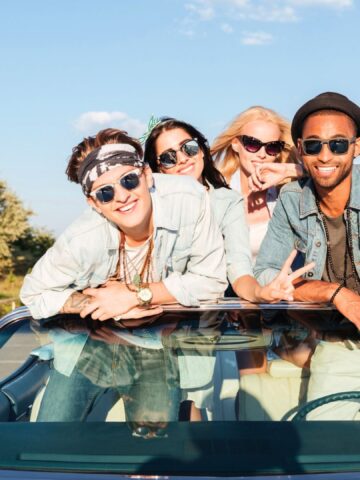 Four young friends in sunglasses lean out of a convertible car, smiling and posing on a sunny day by the water.