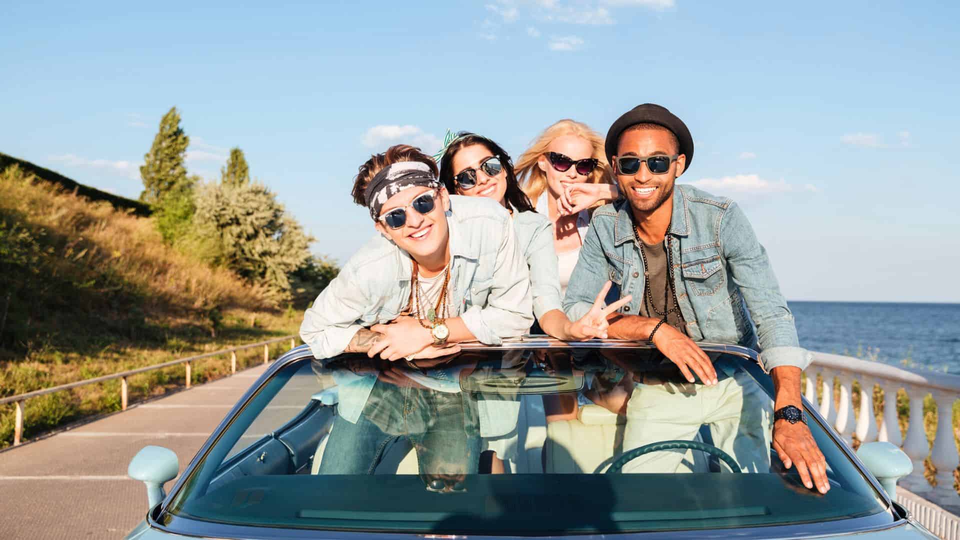 Four young friends in sunglasses lean out of a convertible car, smiling and posing on a sunny day by the water.