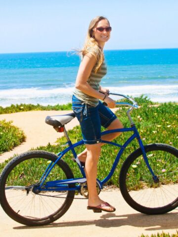 Woman smiling on a blue bicycle near the beach, with ocean and greenery in the background.