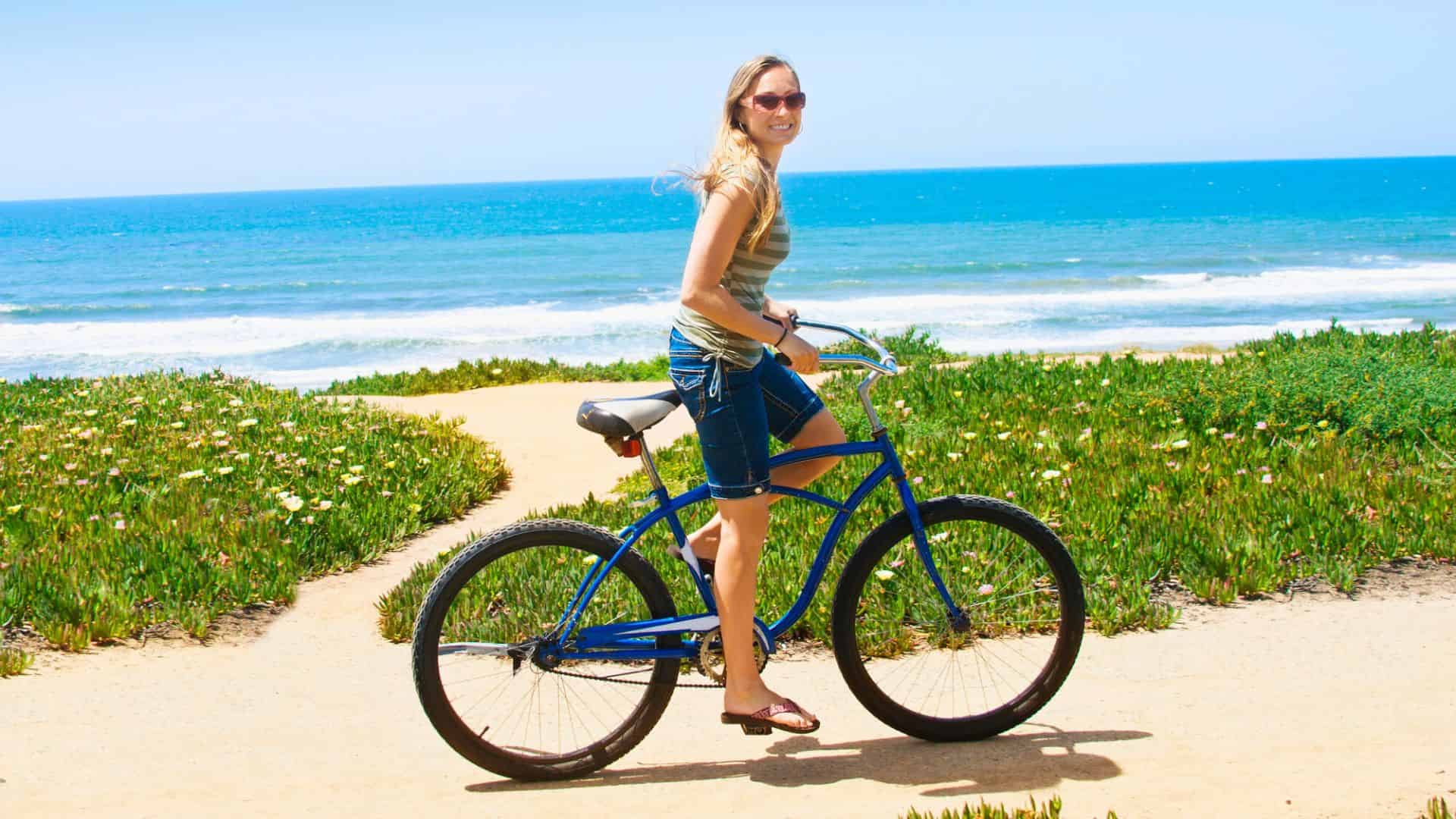 Woman smiling on a blue bicycle near the beach, with ocean and greenery in the background.