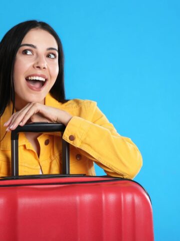 Smiling woman in yellow jacket with red suitcase points up against a bright blue background.