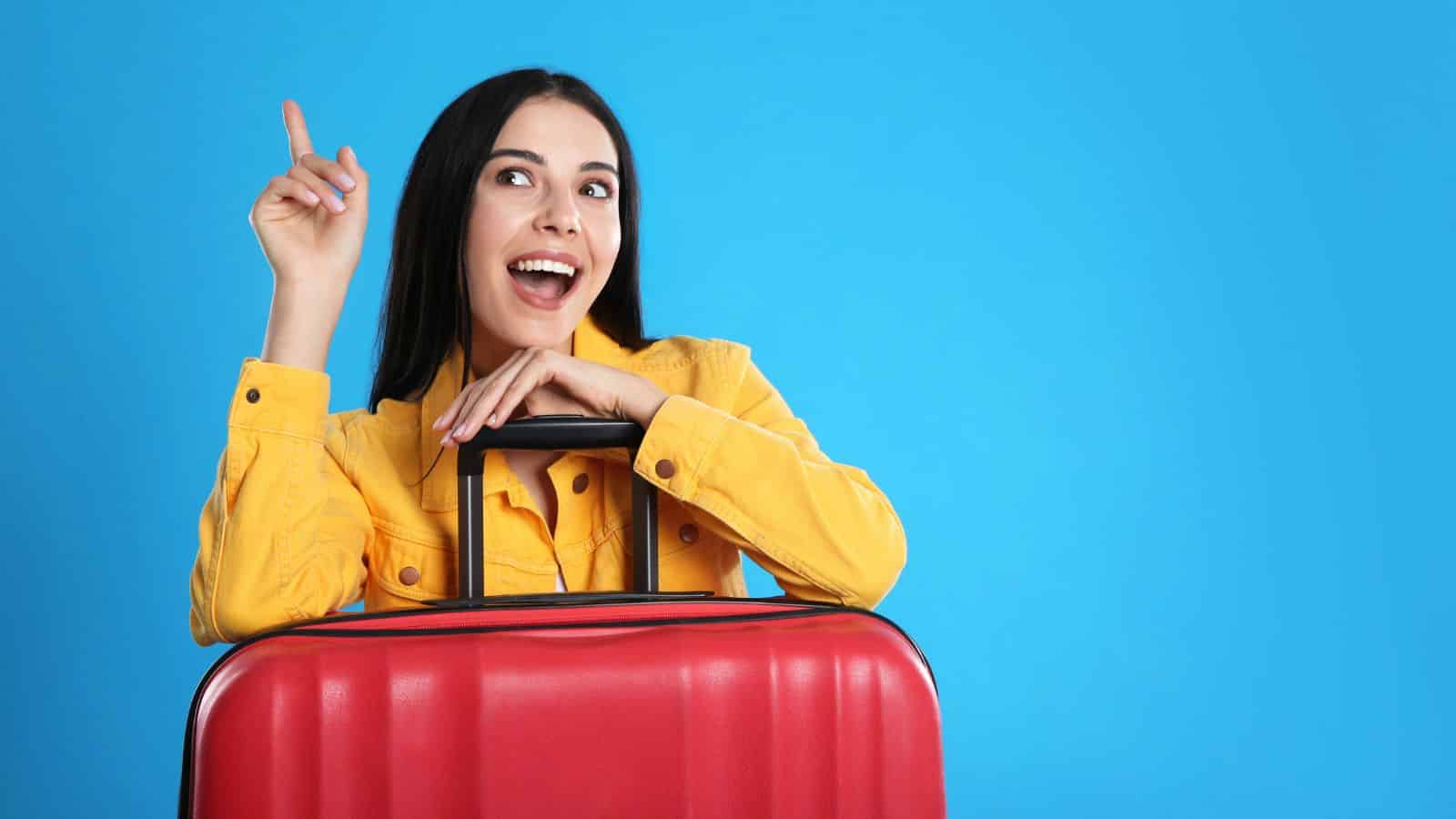 Smiling woman in yellow jacket with red suitcase points up against a bright blue background.