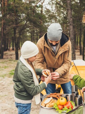 A family enjoys a picnic at a campsite, preparing food together near a tent in the woods.