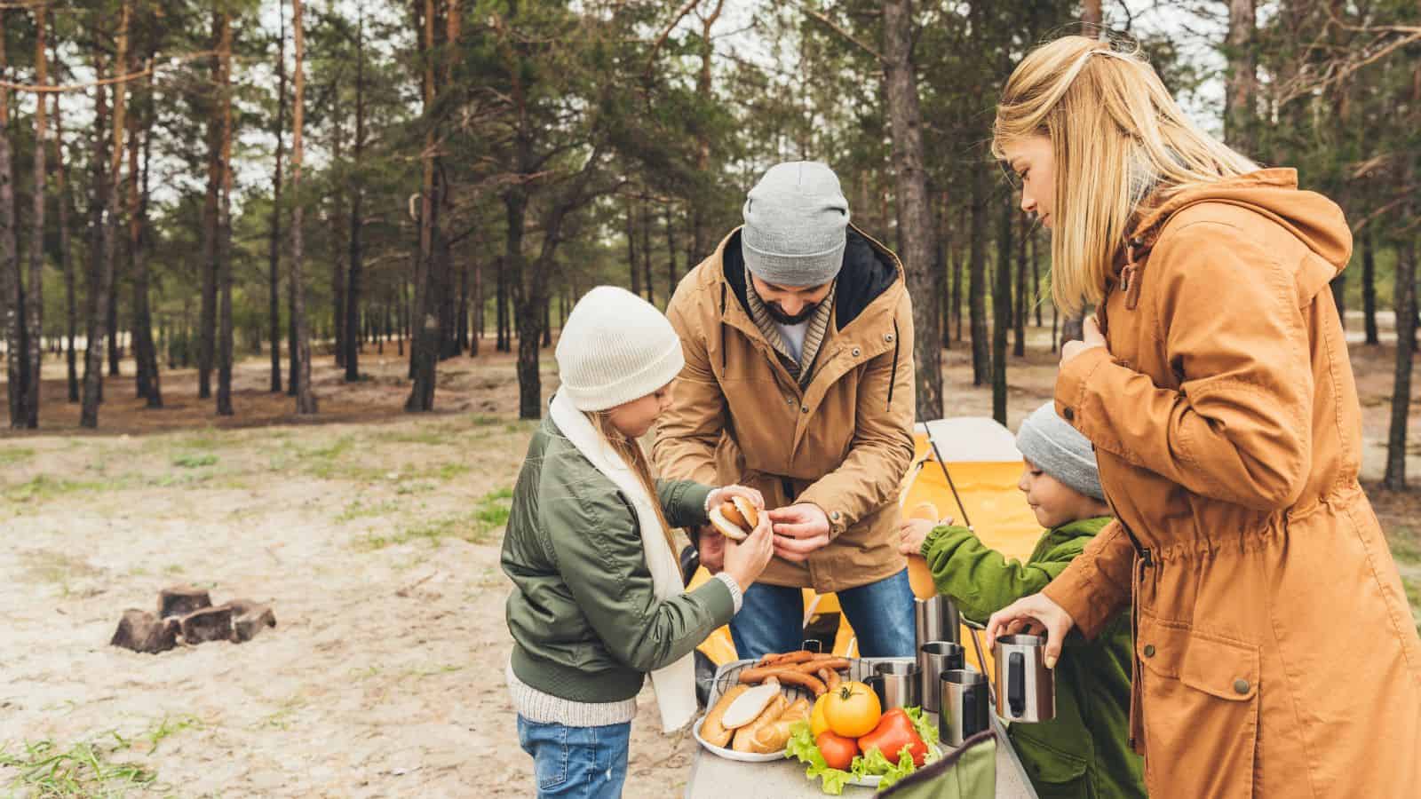 A family enjoys a picnic at a campsite, preparing food together near a tent in the woods.