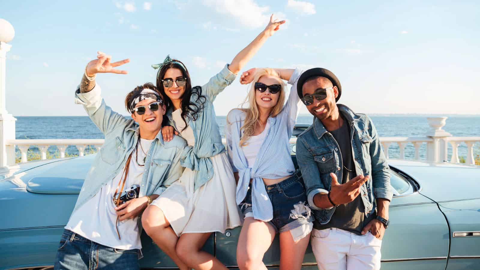 Four friends in casual summer clothes pose and smile by a blue car near the ocean on a sunny day.