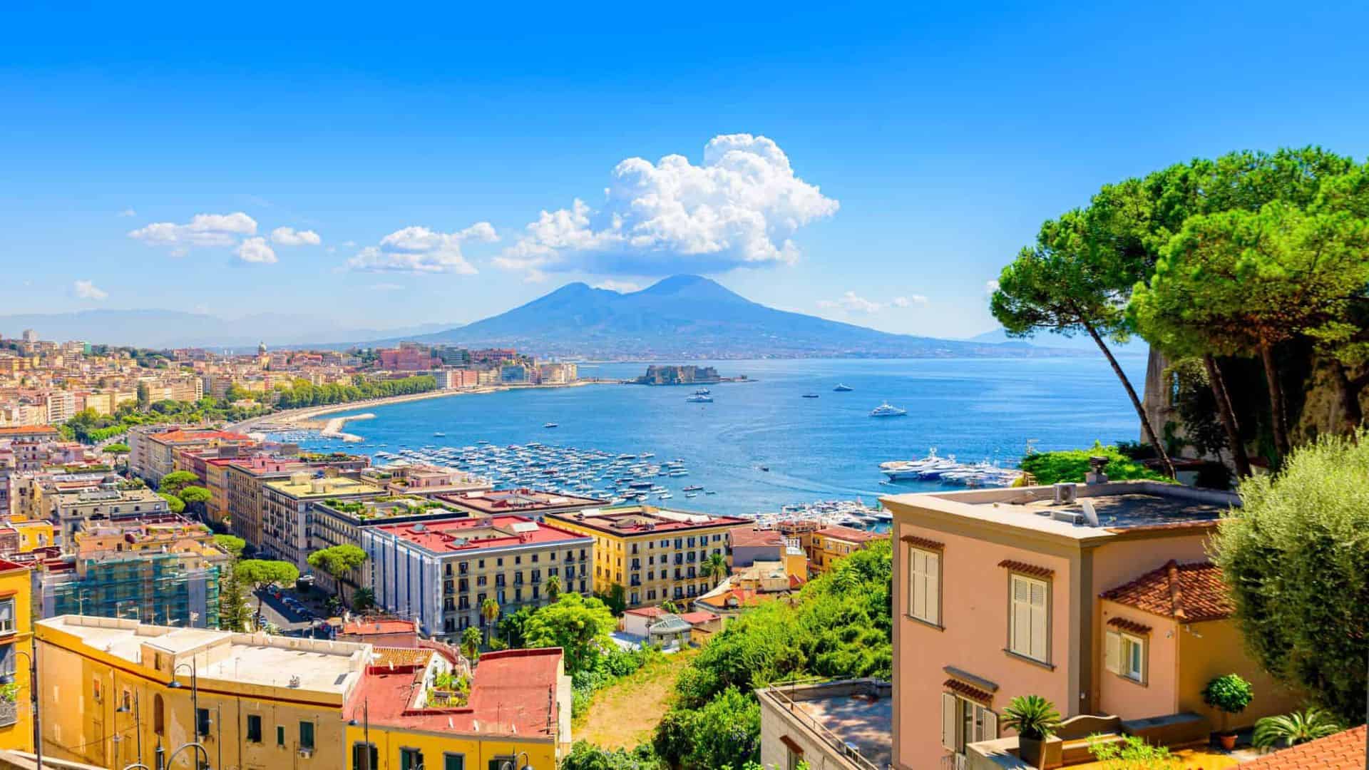 Colorful view of Naples, Italy, with Mount Vesuvius and the bay under a bright blue sky with scattered clouds.