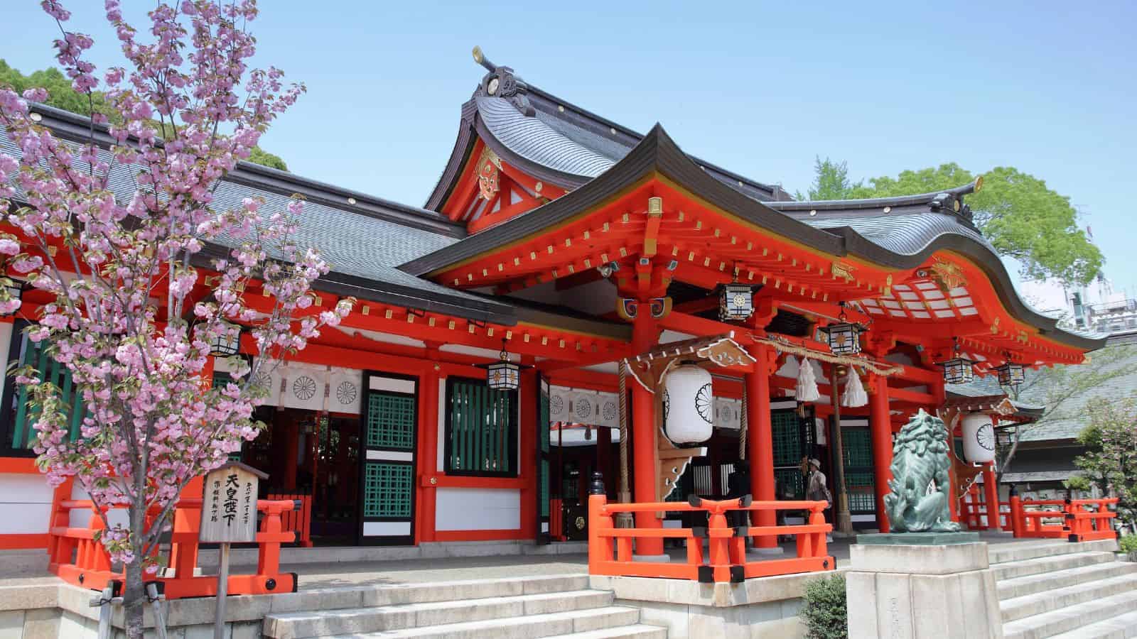 Traditional Japanese shrine with red pillars, ornate roof, lanterns, and blooming cherry blossom tree in front.