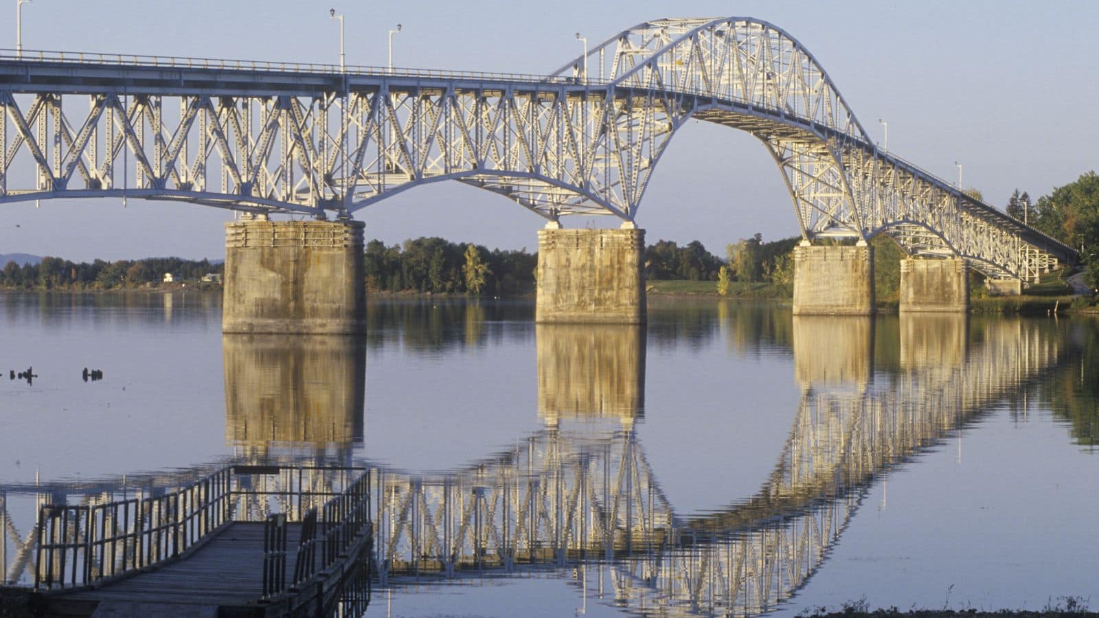 Large steel bridge with arches spans calm river, reflecting on the water under a clear sky.