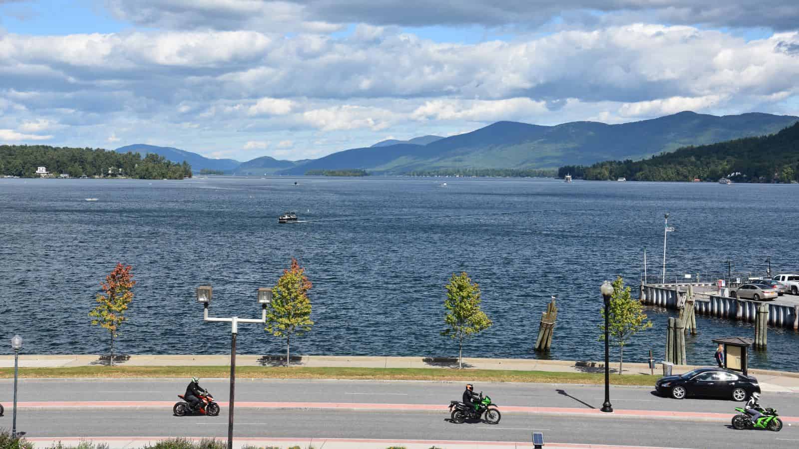 Motorcyclists ride by a lakeside road with mountains and boats in the background under a partly cloudy sky.