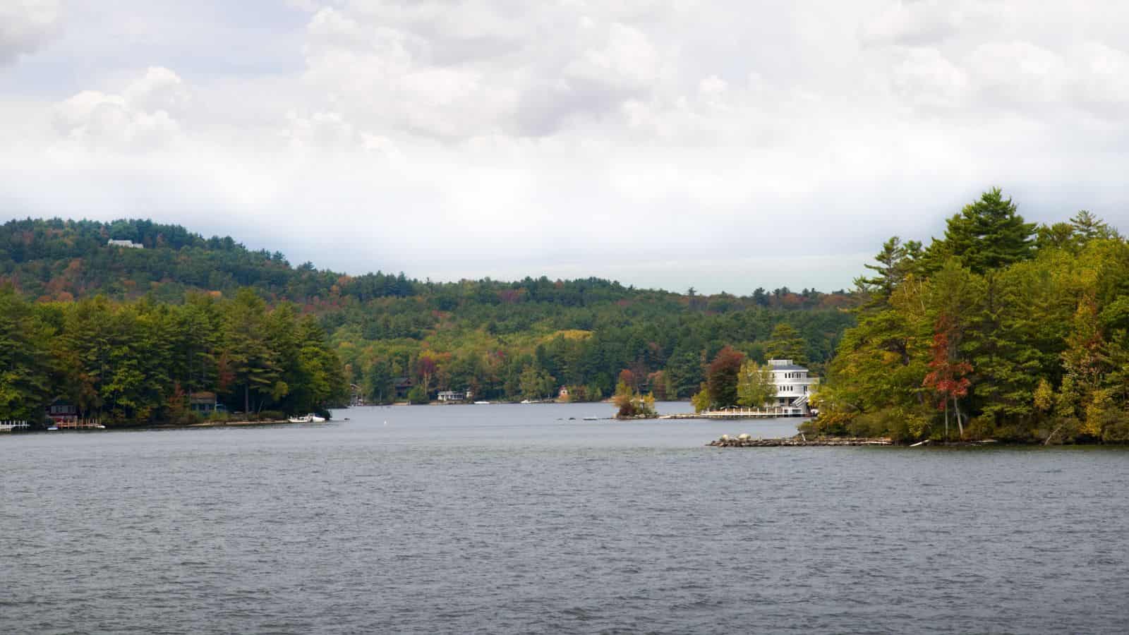 Calm lake with forested hills and scattered houses on the shoreline under a cloudy sky.