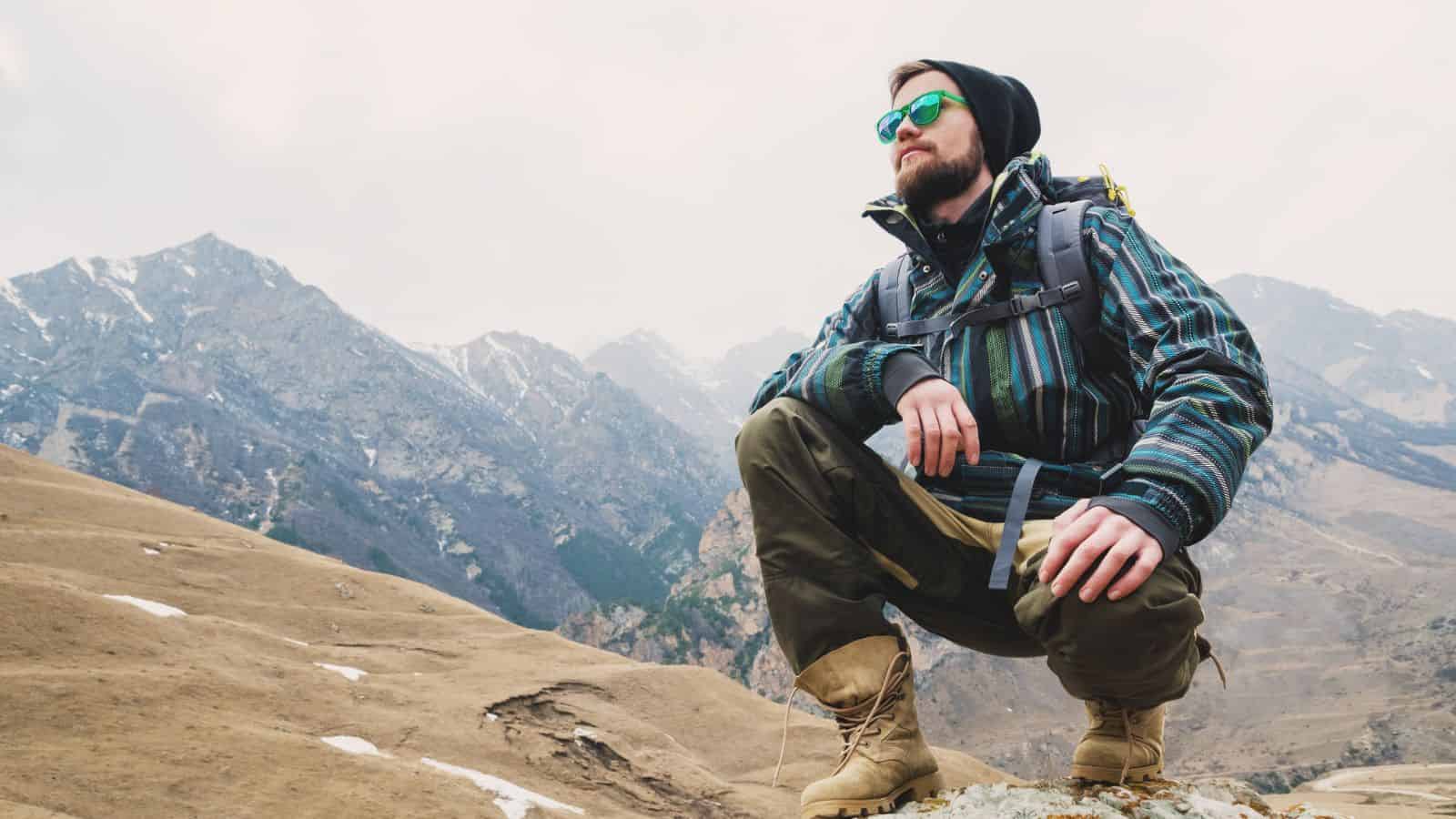 Man wearing sunglasses and hiking gear squats on a rocky hill with mountains in the background.