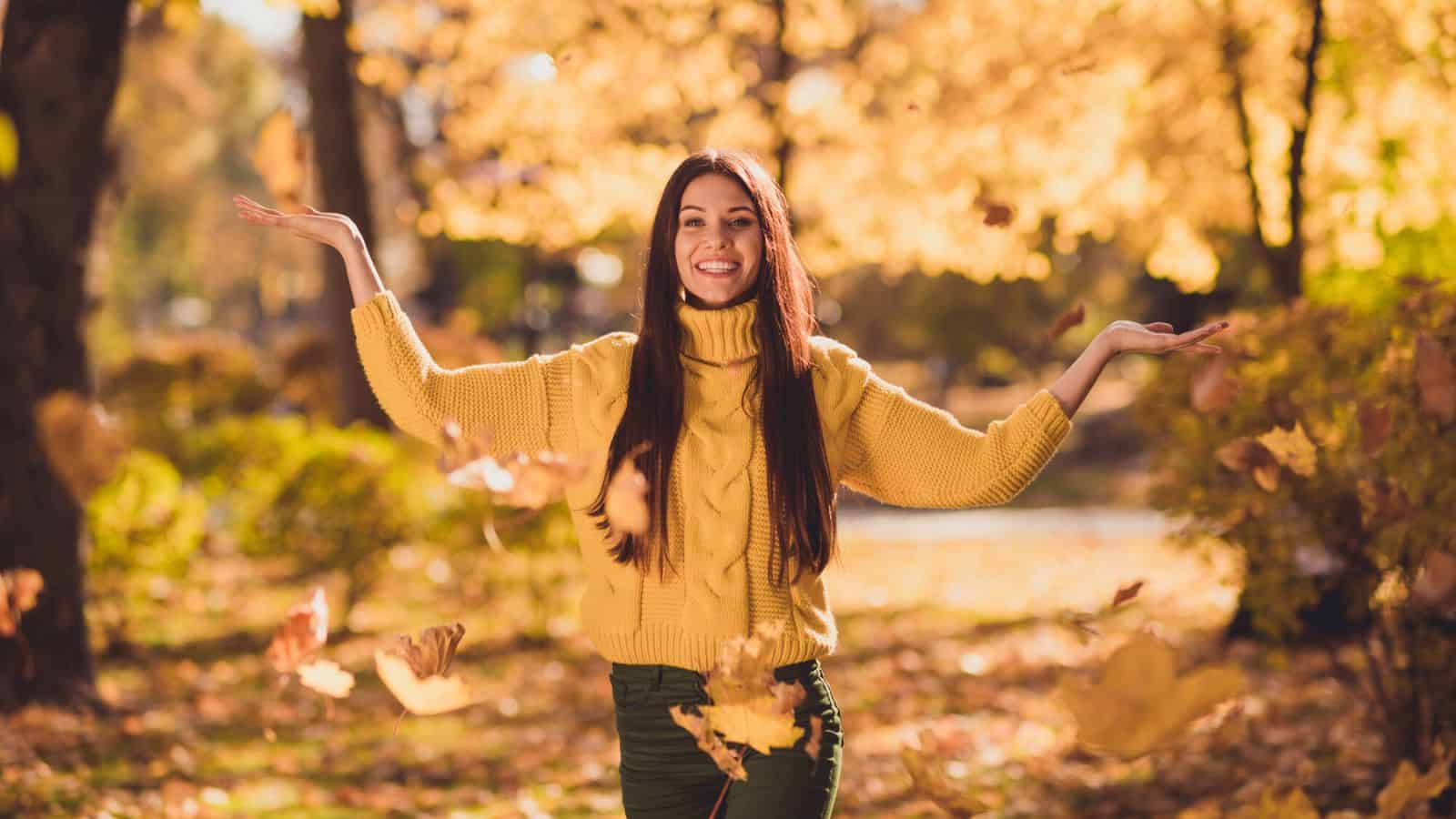 Woman in yellow sweater smiling with arms raised, standing outdoors among falling autumn leaves.