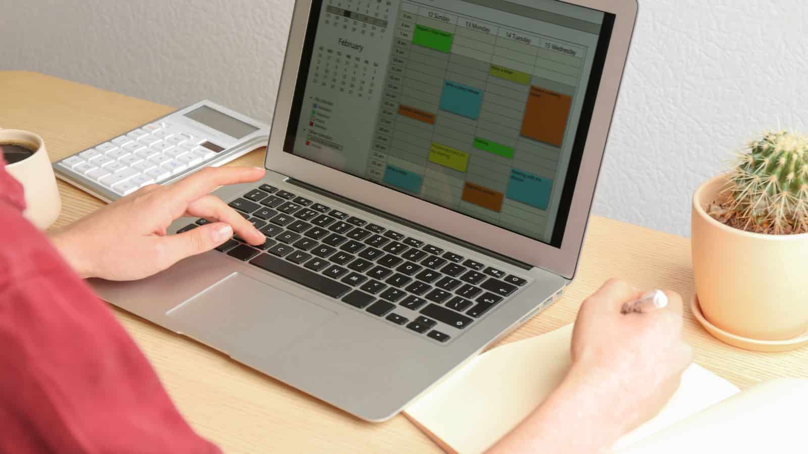 Person using a laptop with a colorful calendar on screen, writing in a notebook, desk with cactus and calculator.