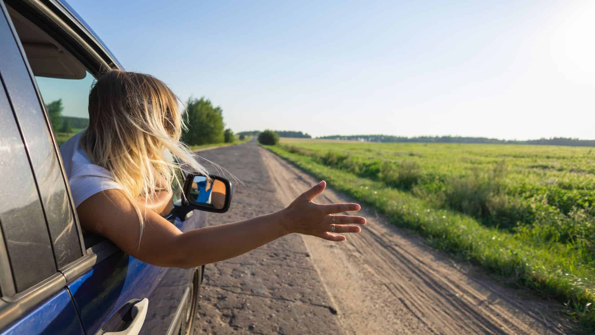 Person leans out of car window, arm outstretched toward a sunny rural road and green fields.