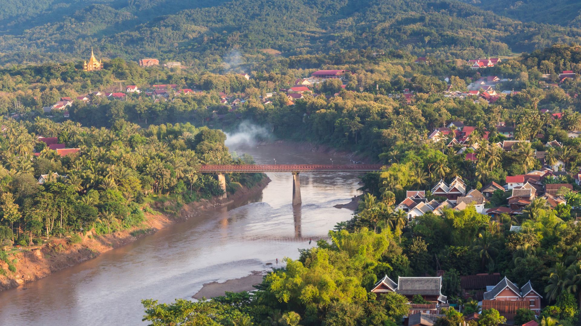 A bridge crosses a river surrounded by trees, houses, and distant forested hills under a clear sky.