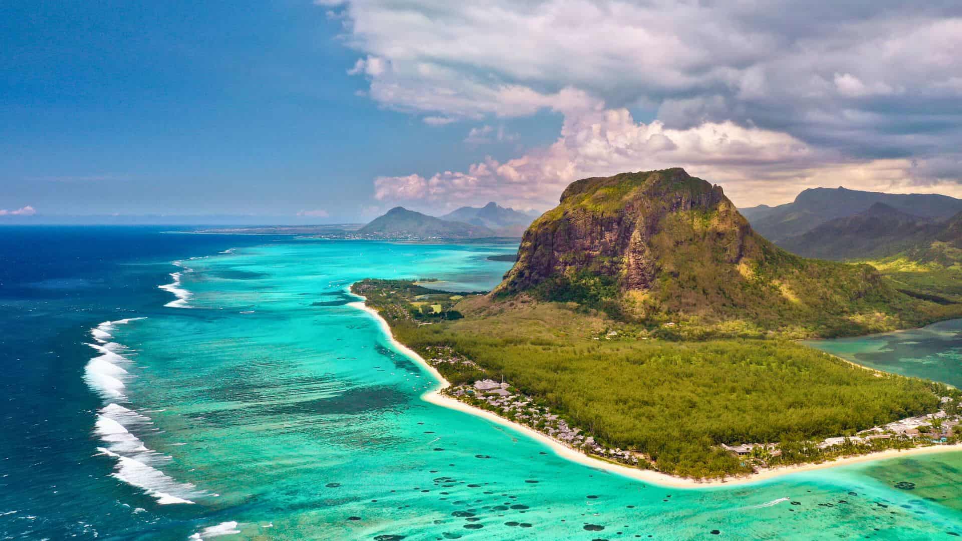 Aerial view of a turquoise lagoon, white sandy beach, and a large green mountain under a partly cloudy sky.