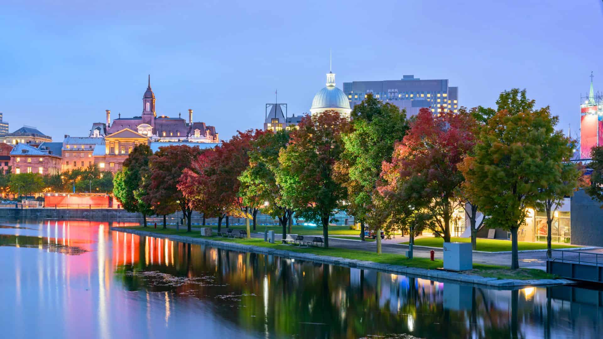 Colorful autumn trees line a waterfront in a city, with historic buildings and lights reflecting in the water.