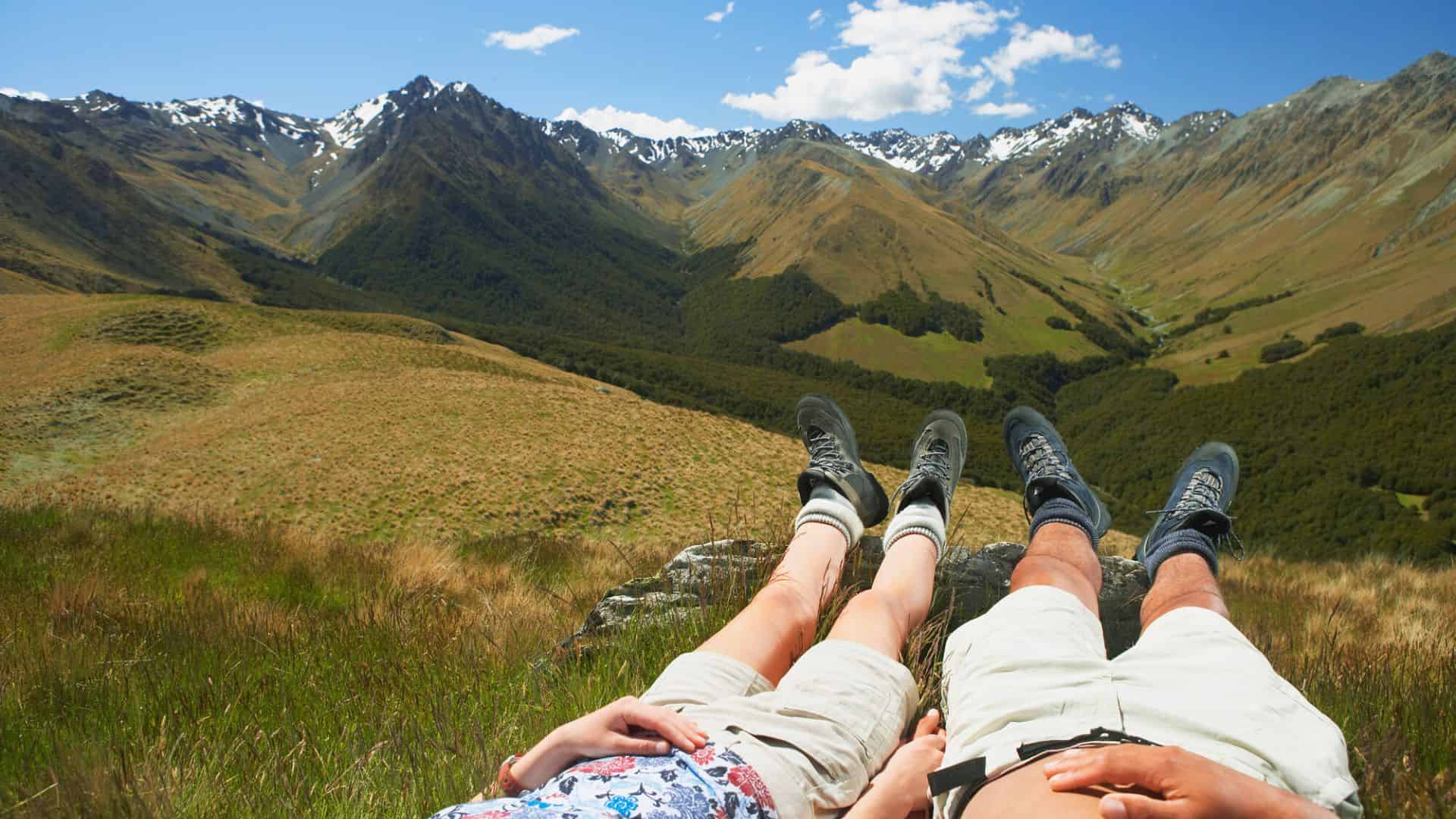 Two people relaxing on grass in front of scenic mountains under a blue sky with scattered clouds.