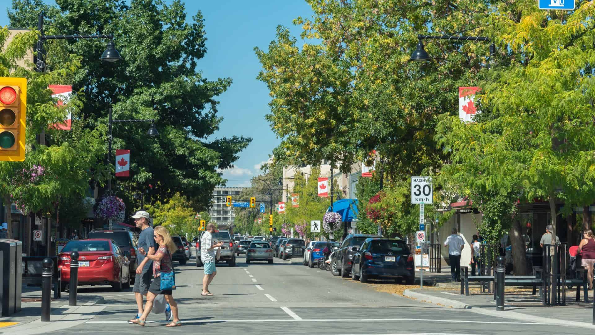 People crossing a tree-lined street decorated with Canadian flags on a sunny day, with cars parked on both sides.