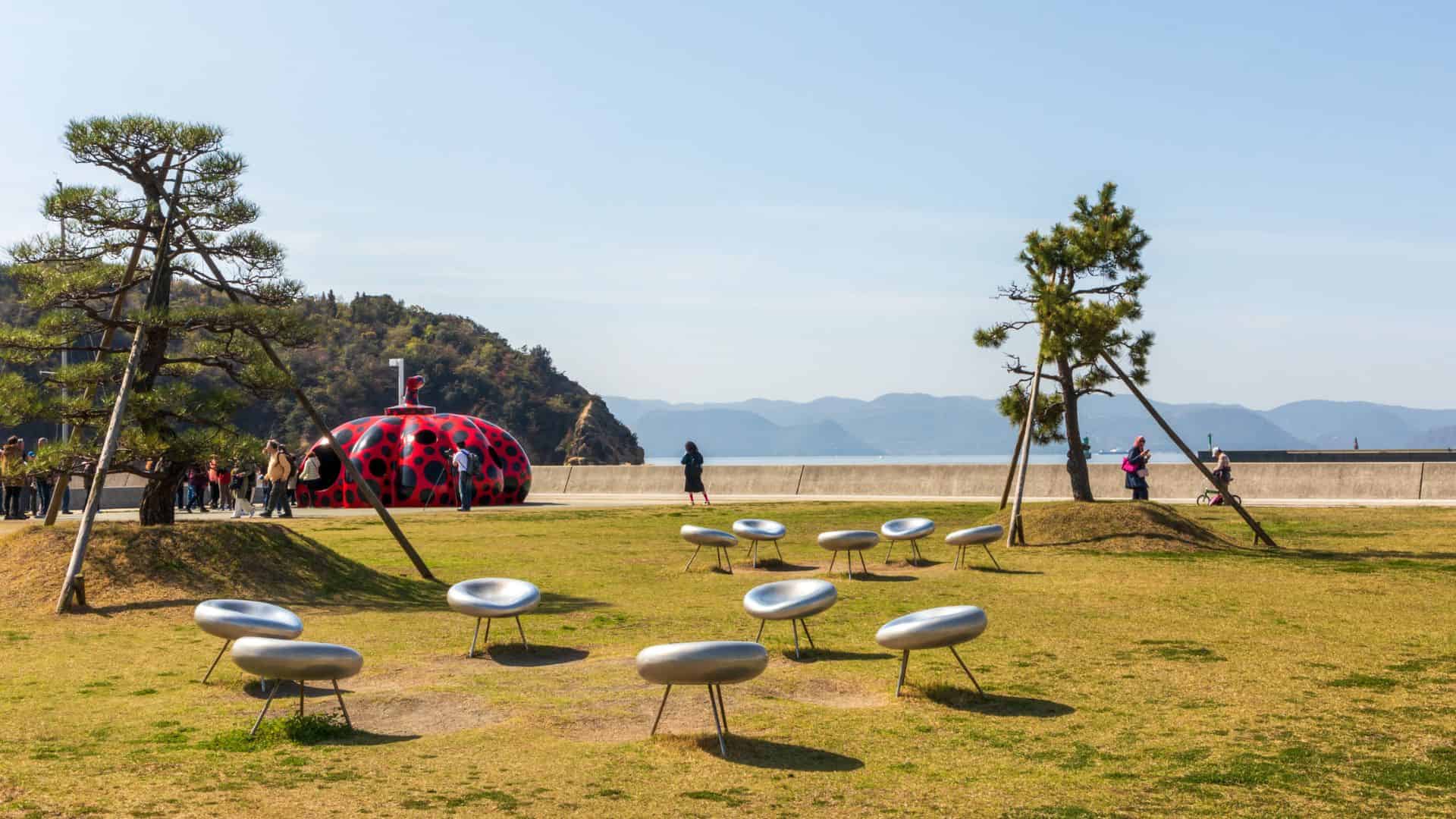 Art installation with silver ring-shaped seats and a red polka-dot sculpture near the sea, with people walking nearby.