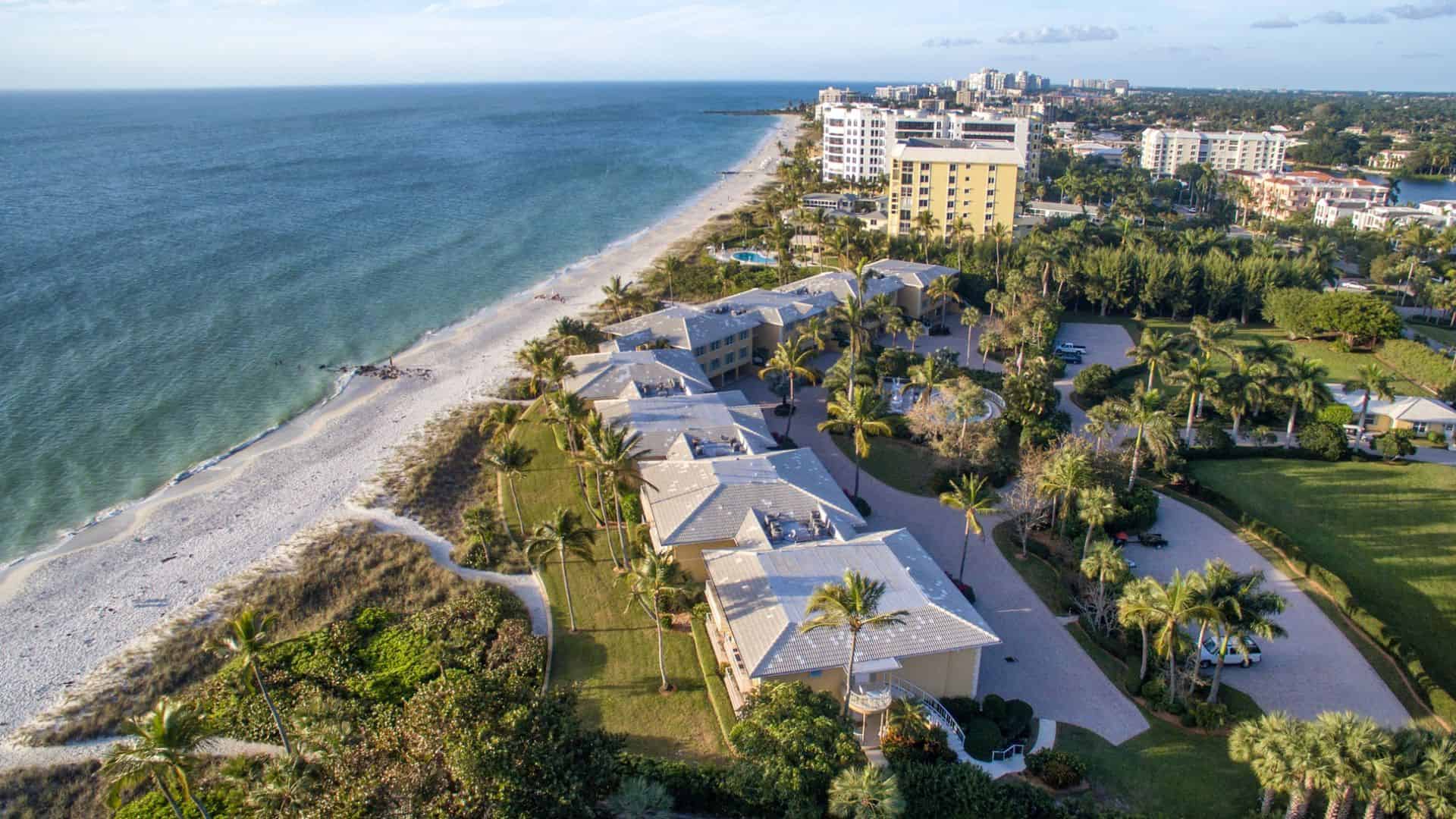 Aerial view of beachfront condos and palm trees along a sandy shoreline with the ocean and city buildings nearby.