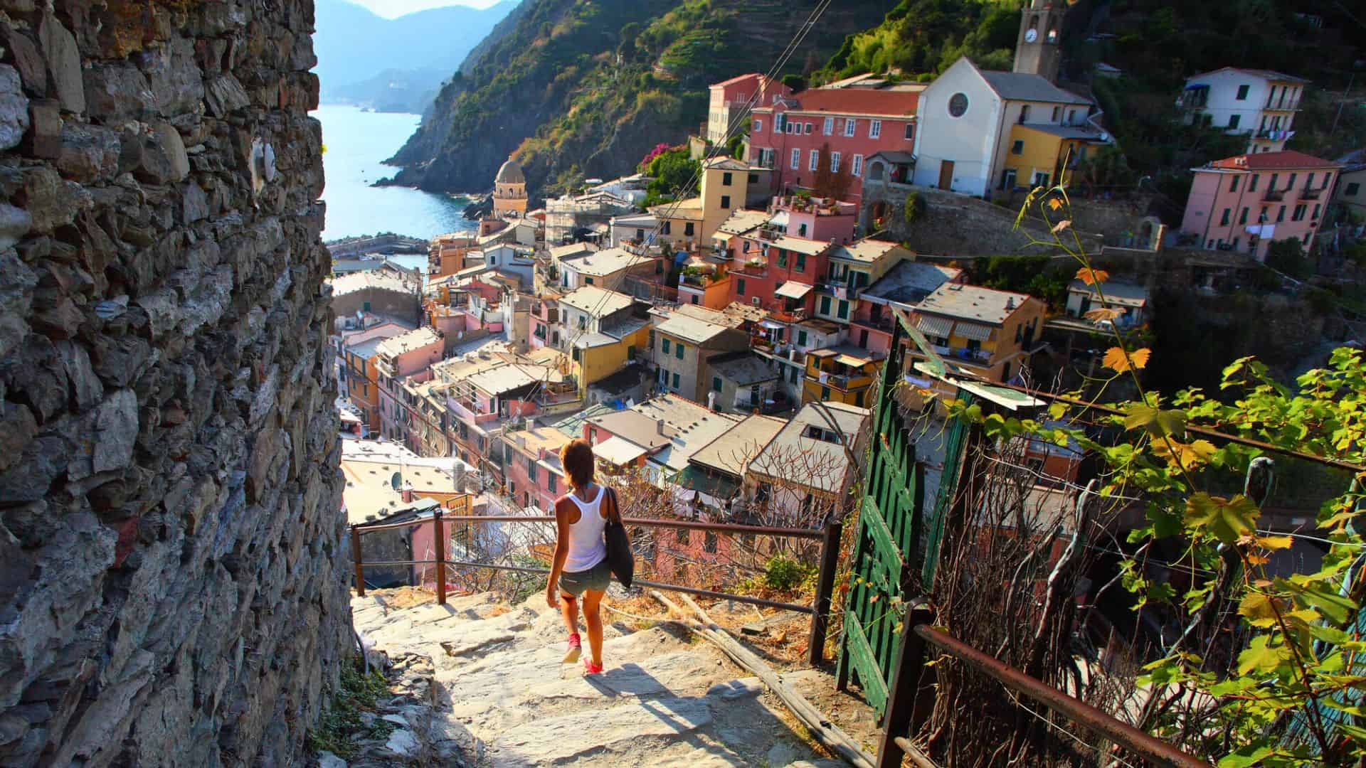 A person walks down stone steps toward colorful coastal houses in a sunny hillside village by the sea.