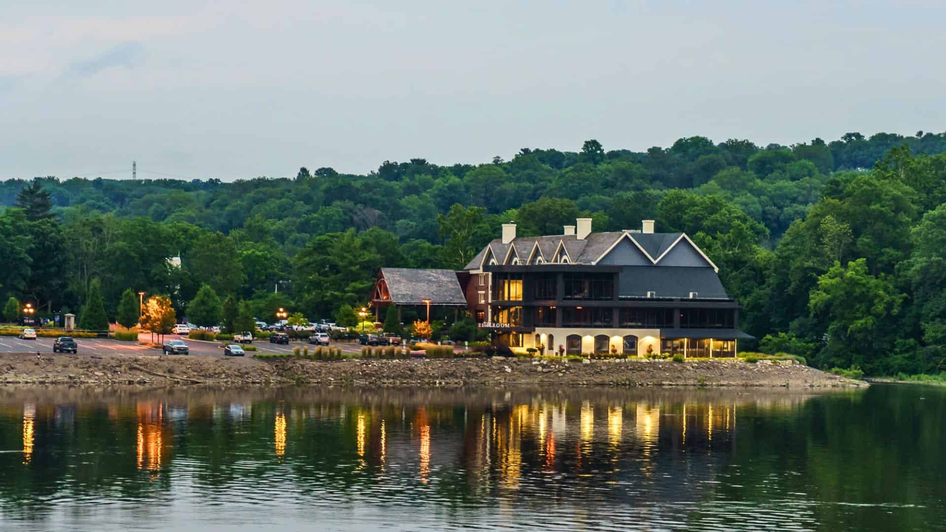 Large building with lights reflecting on calm water, surrounded by trees and parked cars at dusk.