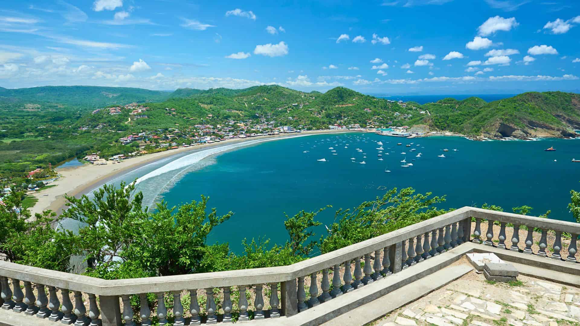 View from a stone balcony overlooking a curving bay with boats, sandy beach, and green hills under a blue sky.