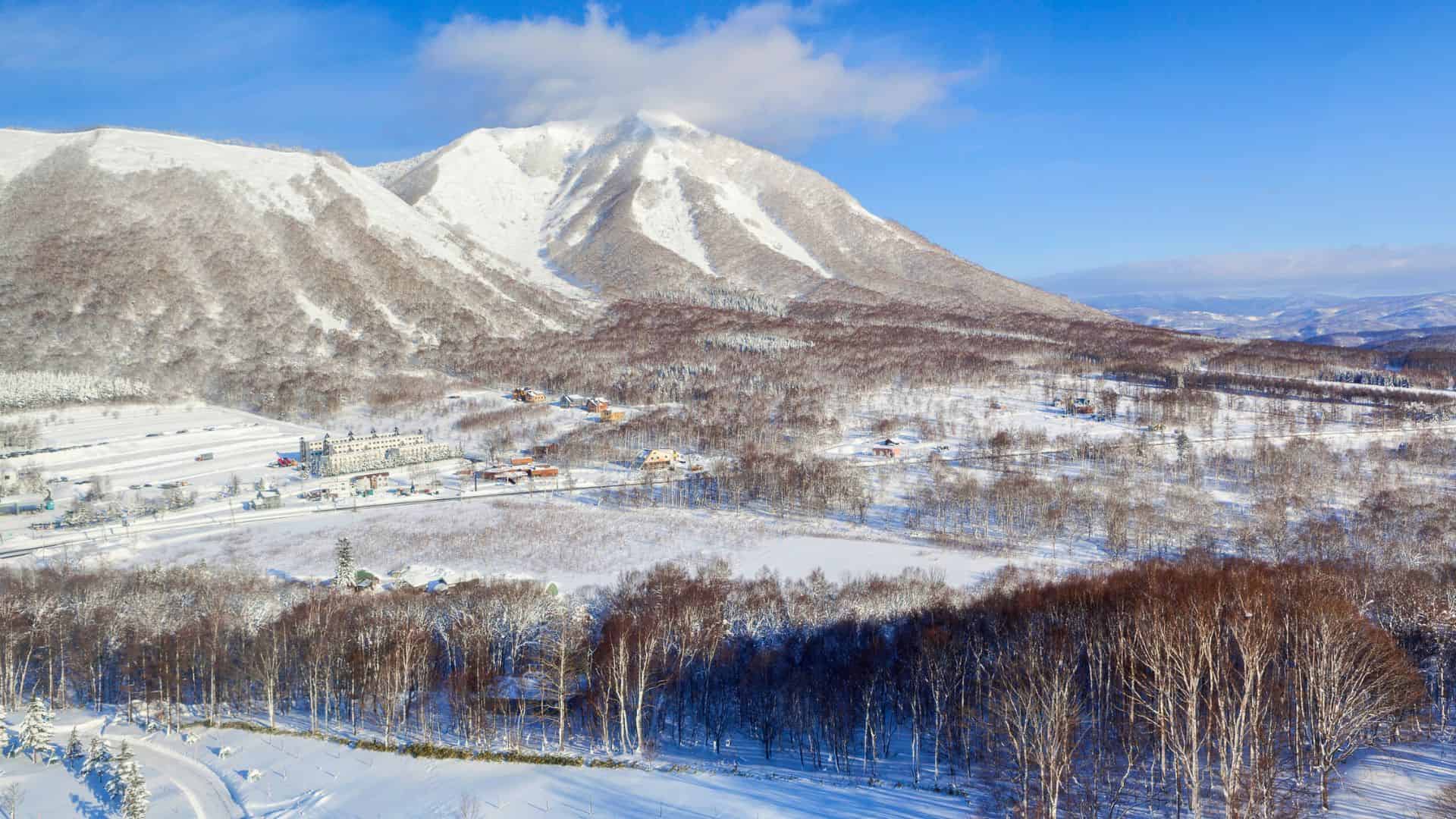 Snowy mountain landscape with scattered trees and buildings under a clear blue sky on a sunny winter day.