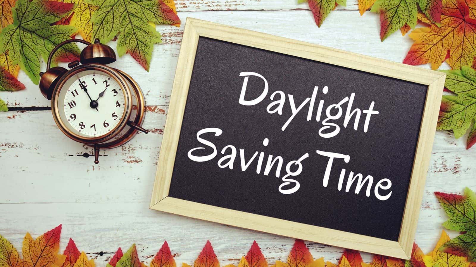 Alarm clock and chalkboard with "Daylight Saving Time," surrounded by colorful autumn leaves.