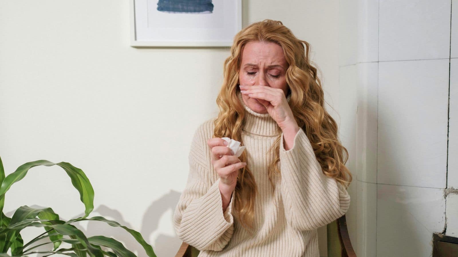 A woman with long hair, in a cream sweater, wipes her nose with a tissue, looking upset.