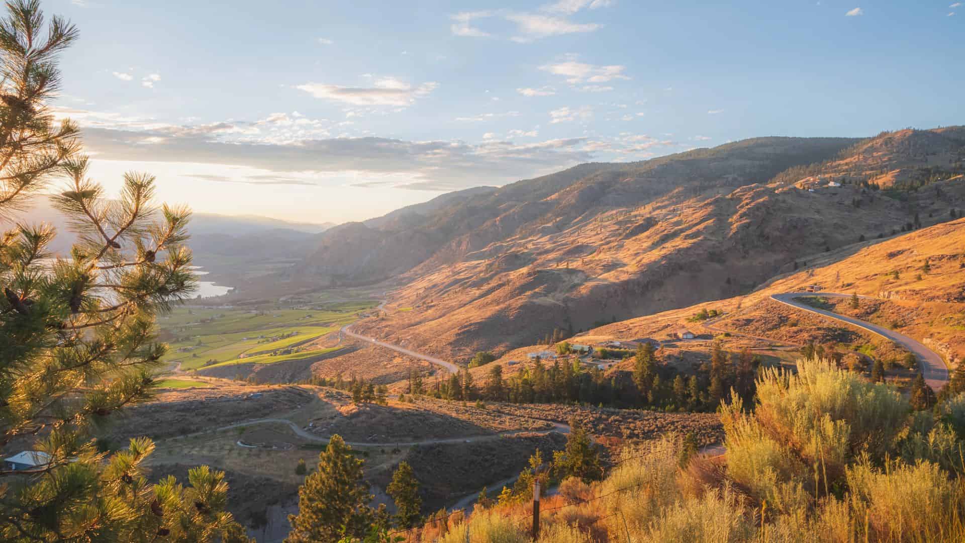 Rolling hills and valleys with winding roads at sunset, with trees and mountains in the background.