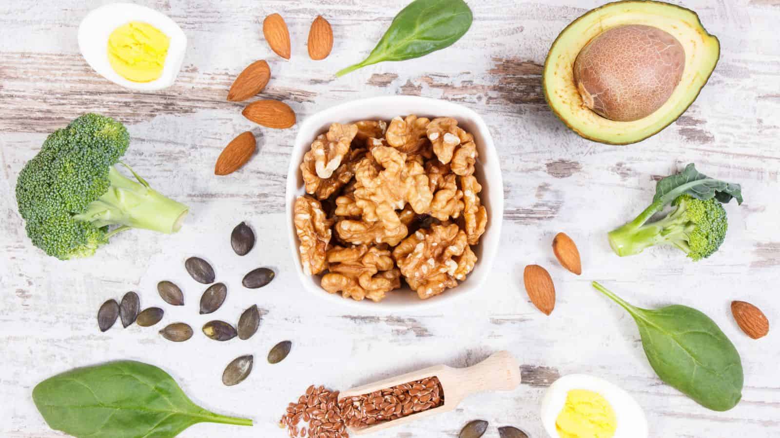A bowl of walnuts surrounded by avocado, broccoli, eggs, almonds, spinach, and seeds on a wooden table.