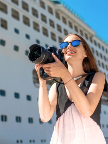 Woman in sunglasses holding a camera, smiling in front of a large white cruise ship on a sunny day.