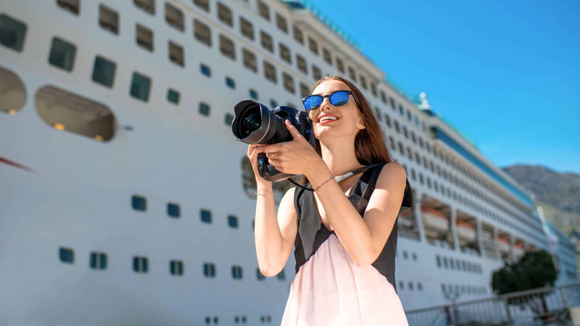 Woman in sunglasses holding a camera, smiling in front of a large white cruise ship on a sunny day.