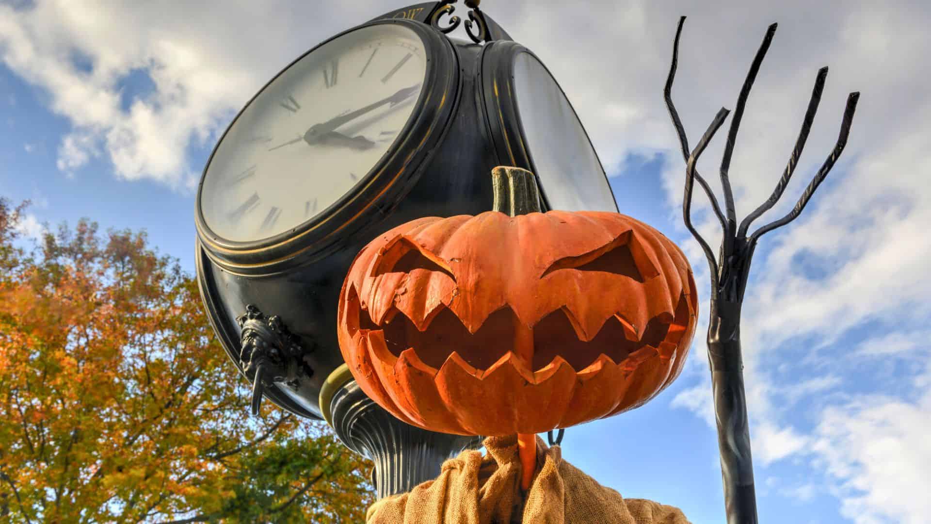 A jack-o’-lantern scarecrow stands in front of a vintage clock and a pitchfork against a cloudy sky.