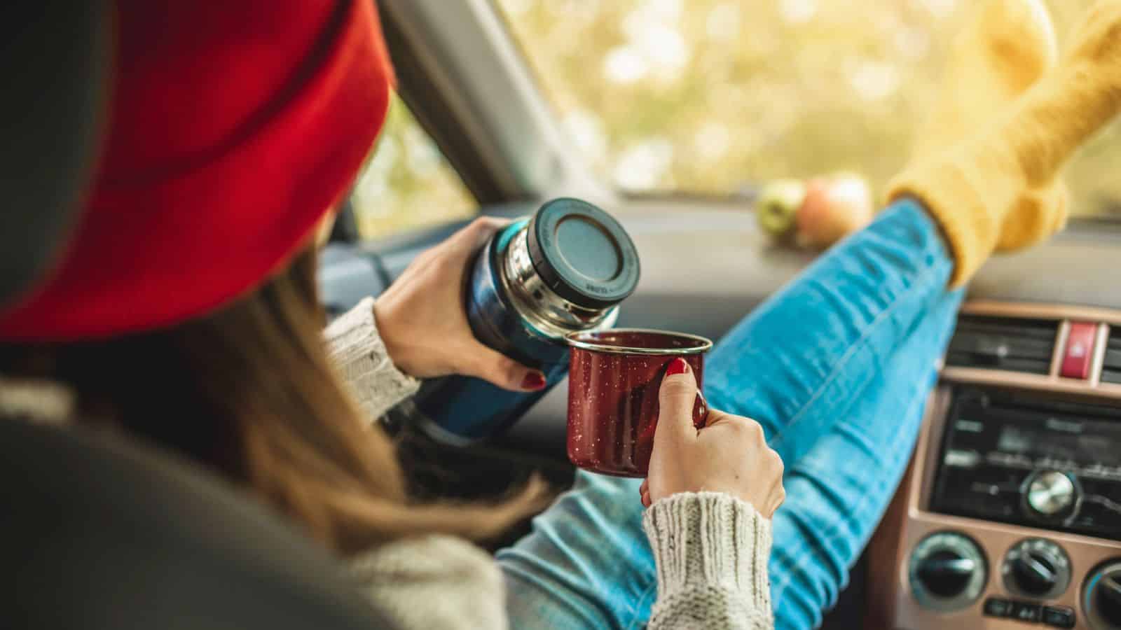 Person in red beanie pours a drink from a thermos into a mug while relaxing in a car with feet on the dashboard.