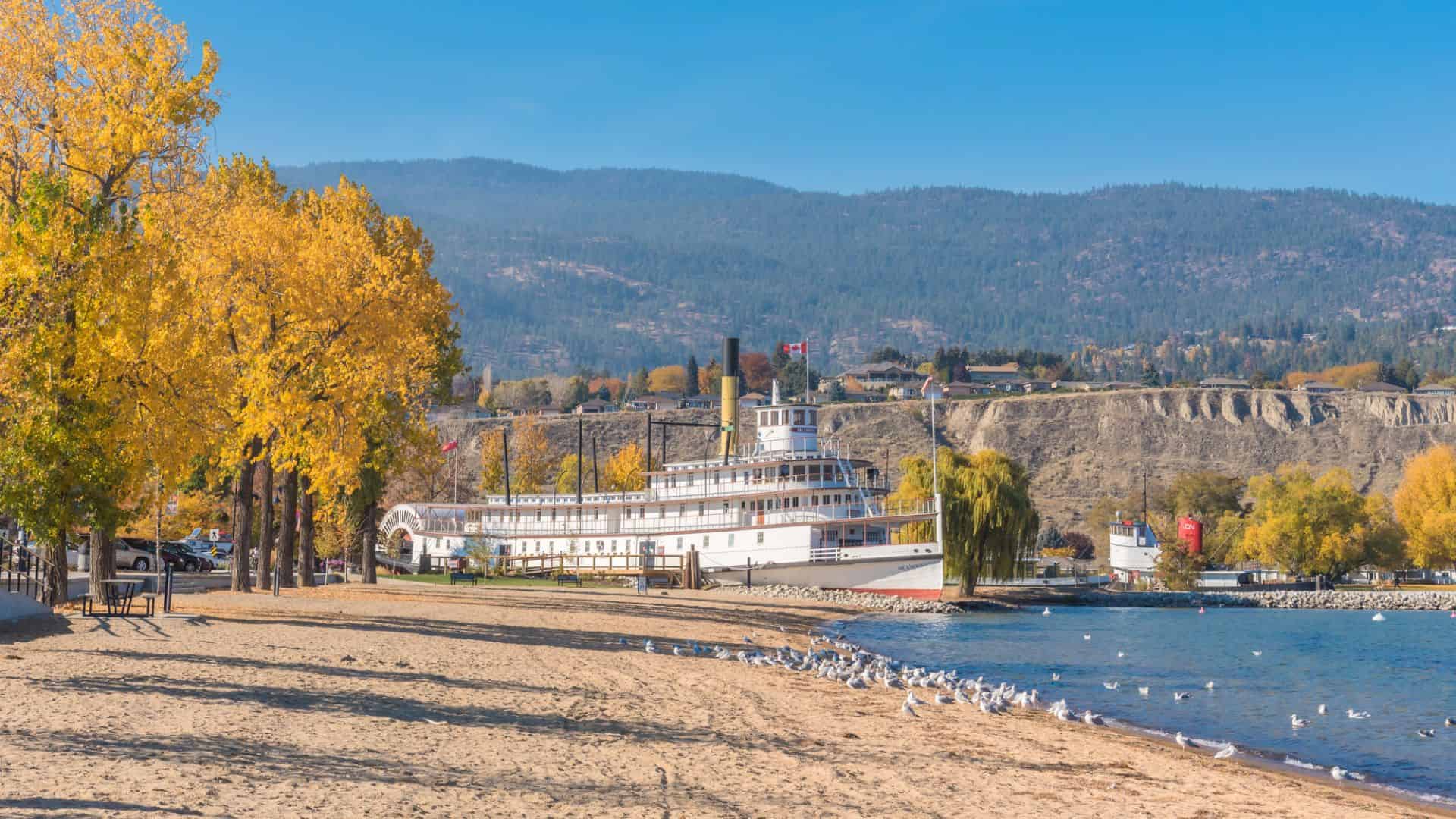 A historic paddle wheeler docked by a sandy beach with autumn trees and distant hills under a blue sky.