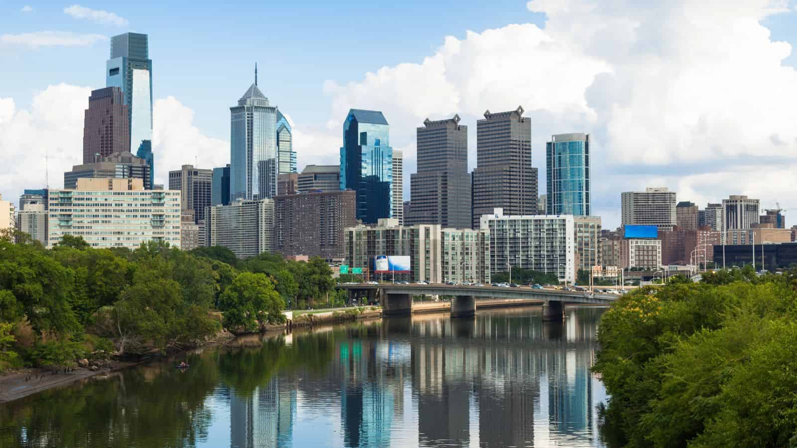 Philadelphia city skyline with modern skyscrapers and a bridge over a river, surrounded by green trees.