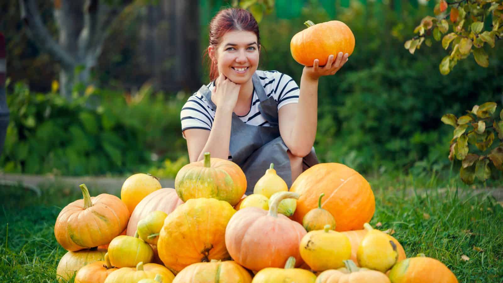 Woman smiling in a garden, holding a pumpkin, surrounded by a pile of various pumpkins.