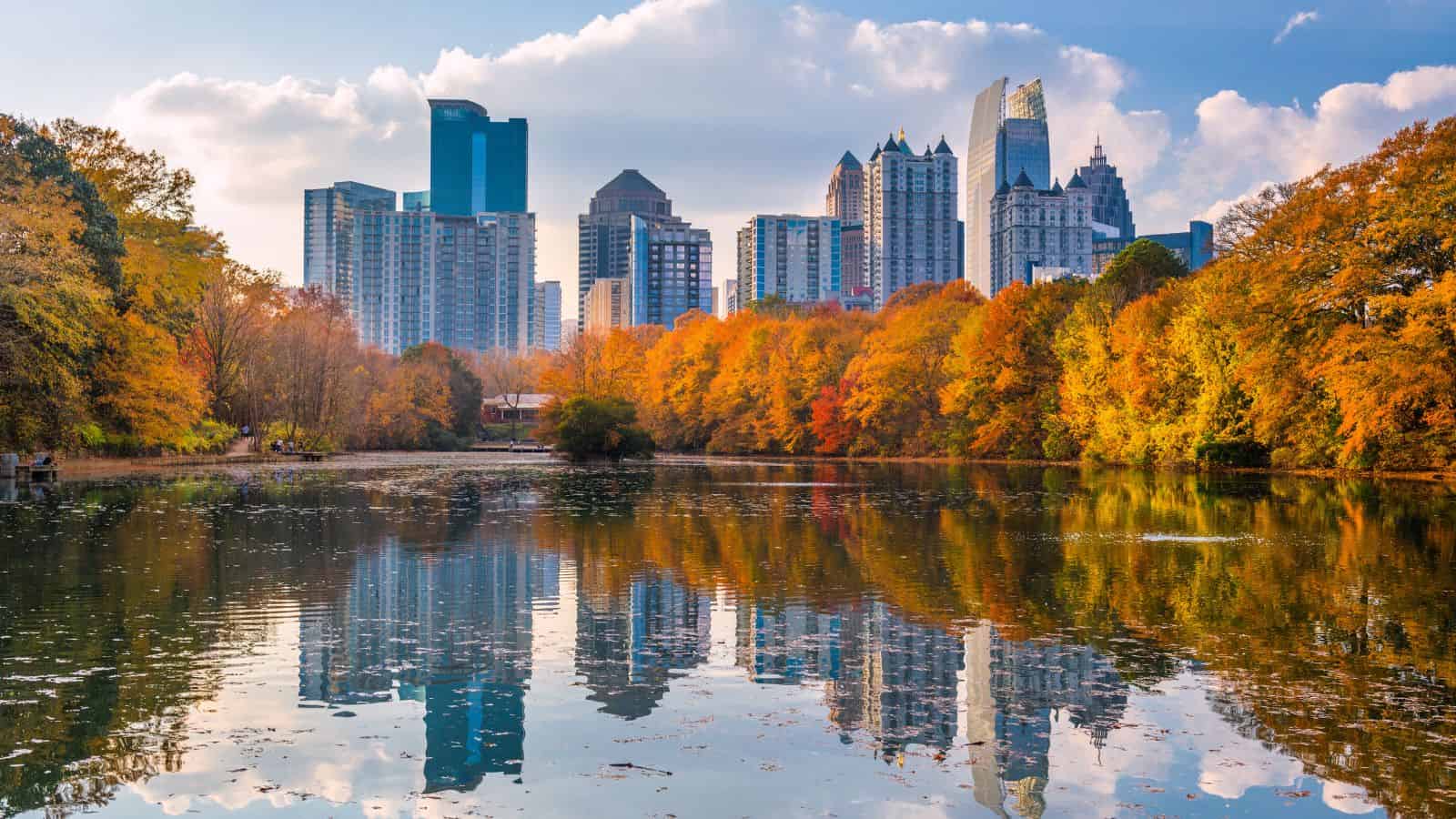 City skyline with modern buildings behind colorful autumn trees reflected in a calm lake under a partly cloudy sky.