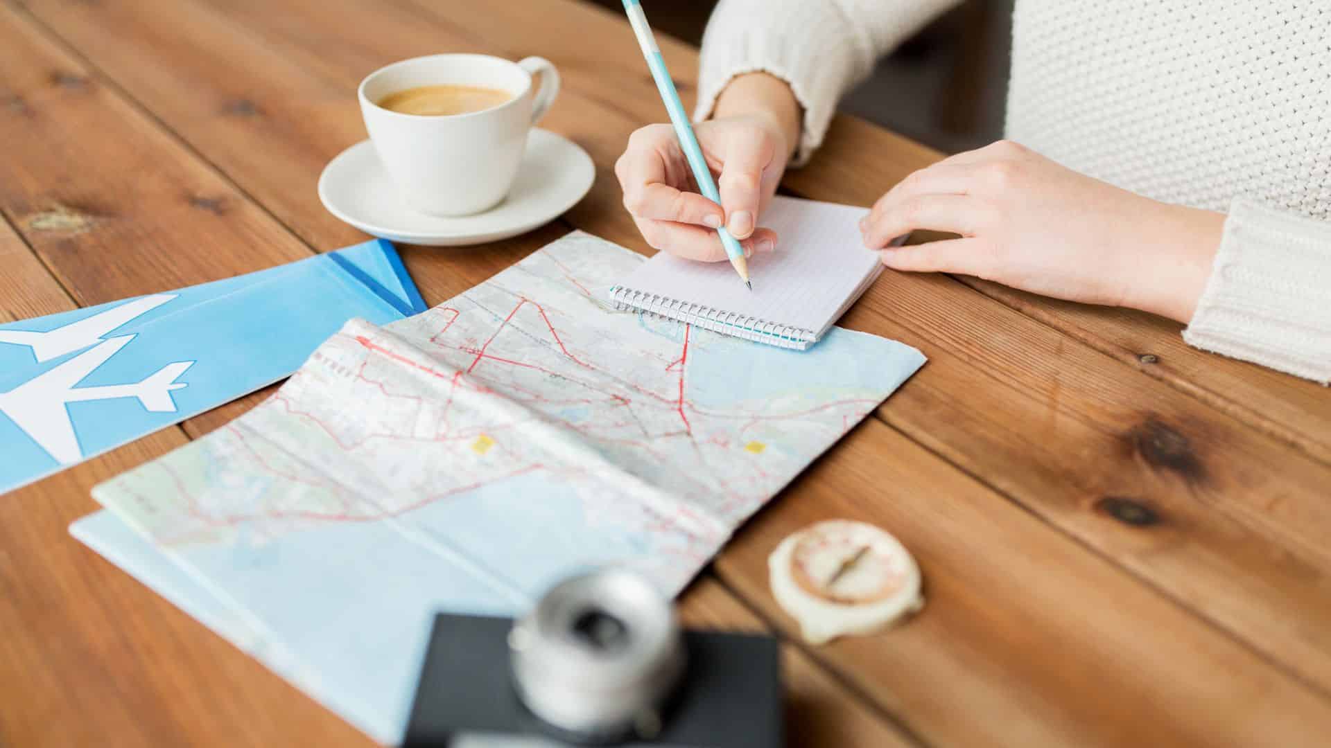 Person writing in a notepad next to a map, compass, camera, coffee, and airline ticket on a wooden table.