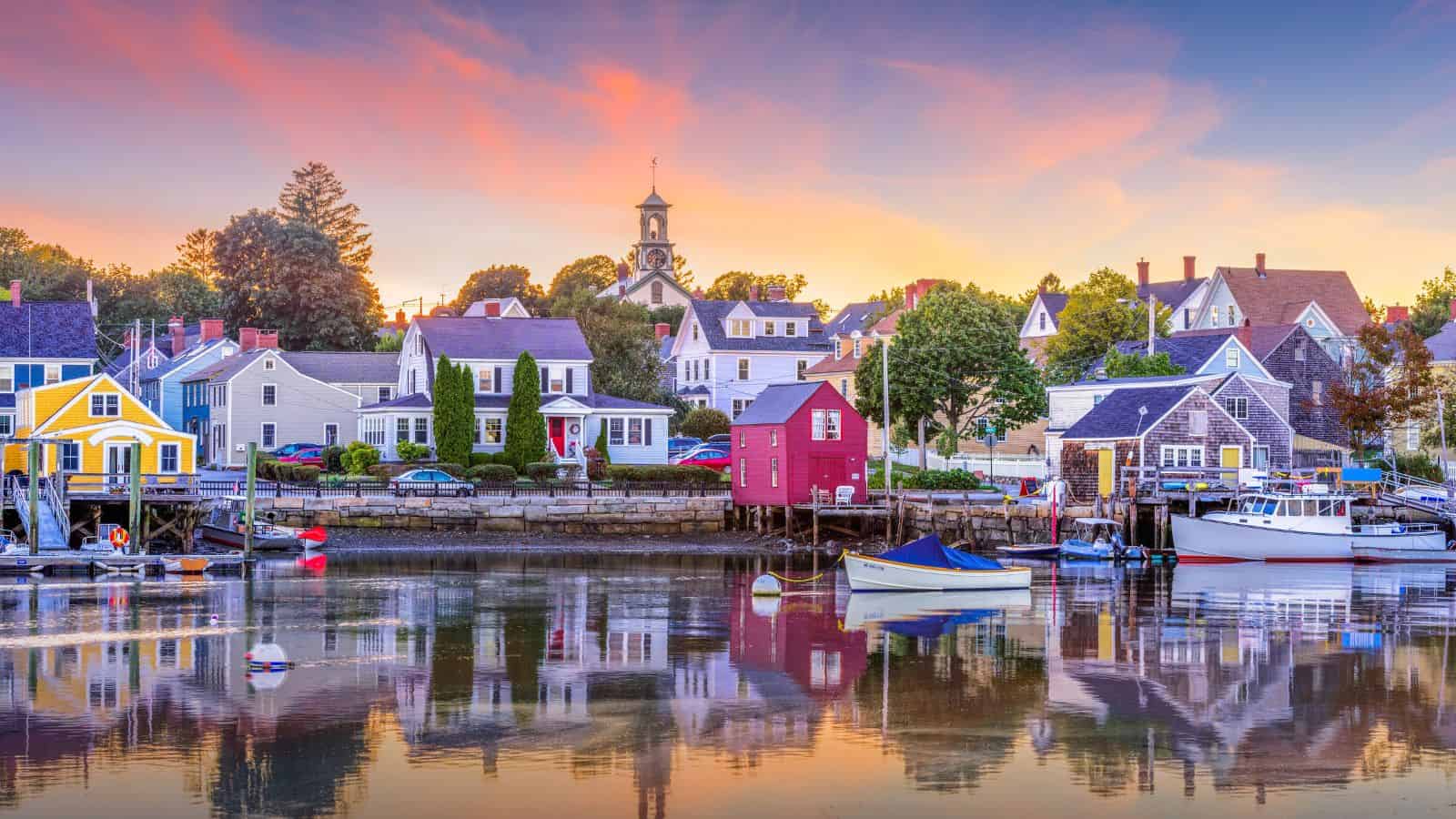 Colorful houses and boats line a calm waterfront at sunset, with reflections in the water and a church steeple behind.