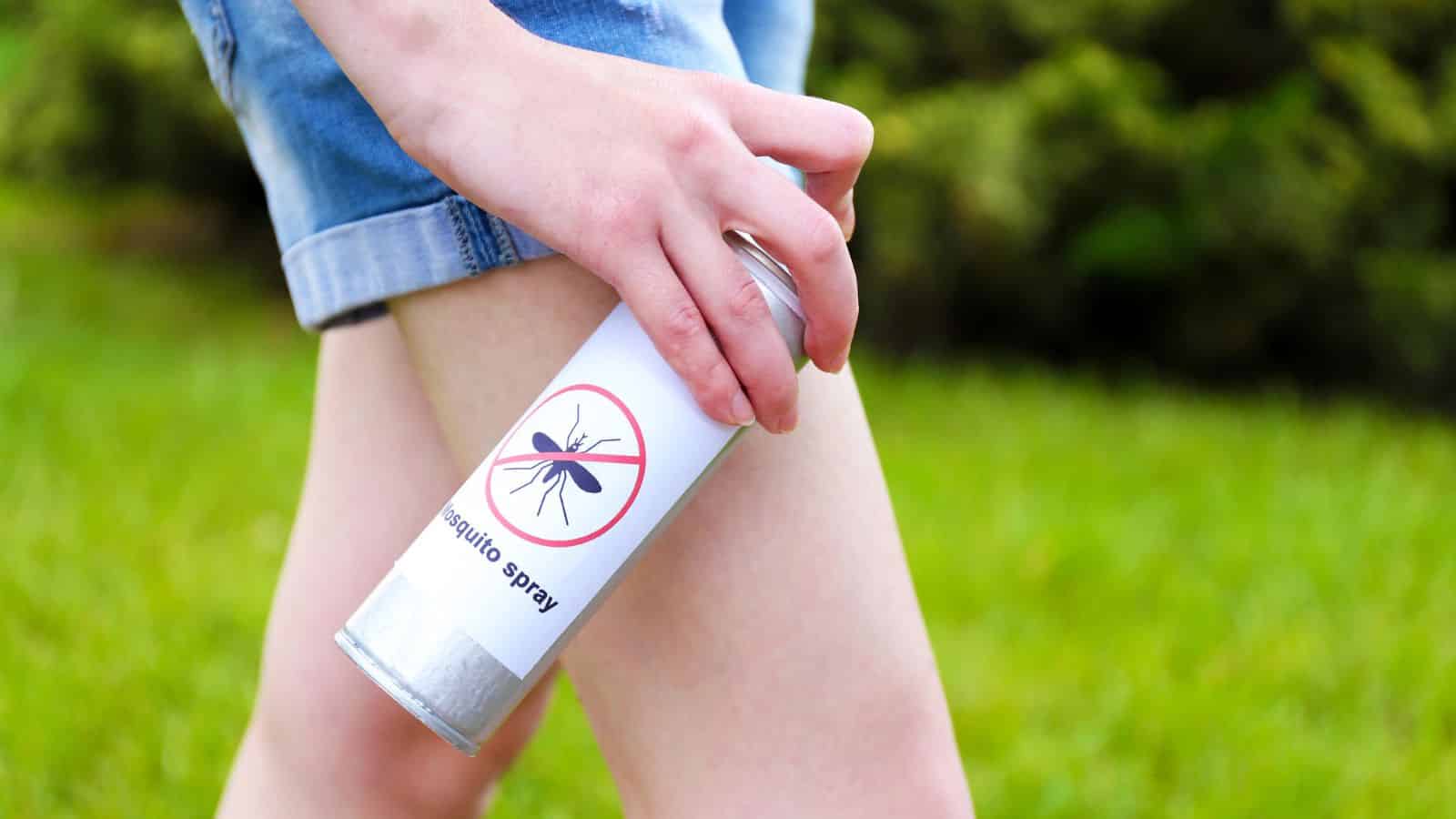 Person spraying mosquito repellent on their leg outdoors, wearing shorts, with grass in the background.