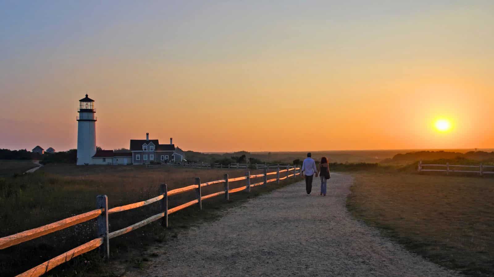 Couple walks along a path toward a lighthouse at sunset, with a wooden fence lining the trail.